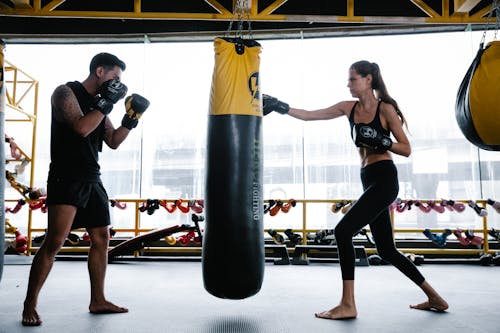 Free Side view of determined fit young female boxer in sportswear and gloves punching bag during workout with male personal trainer standing in stance in modern gym Stock Photo