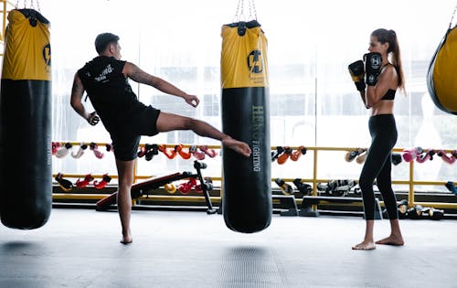 Back view of unrecognizable young muscular male kickboxer hitting punching bag with leg while training in gym with fit female