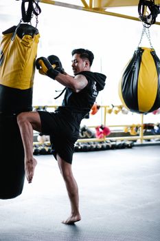 Side view of motivated young Latin American male fighter in sportswear and protective gloves hitting punching bag with leg while practicing kickboxing movements in modern light gym
