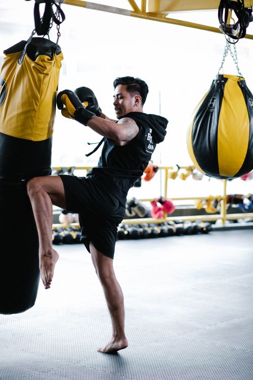 Free Side view of motivated young Latin American male fighter in sportswear and protective gloves hitting punching bag with leg while practicing kickboxing movements in modern light gym Stock Photo