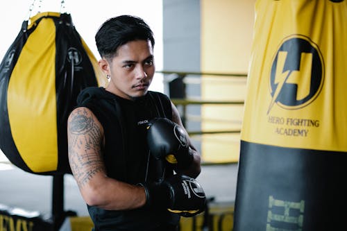 Serious young tattooed Latin American sportsman in boxing gloves looking away attentively while preparing for punching bag in gym