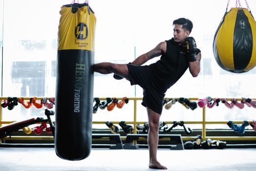 Side view full body of strong Asian sportsman kicking heavy punch bag during workout in gym