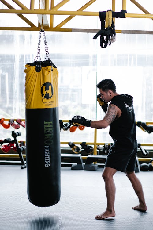 Full length side view of barefoot Asian boxer in gloves punching bag during workout in light gym