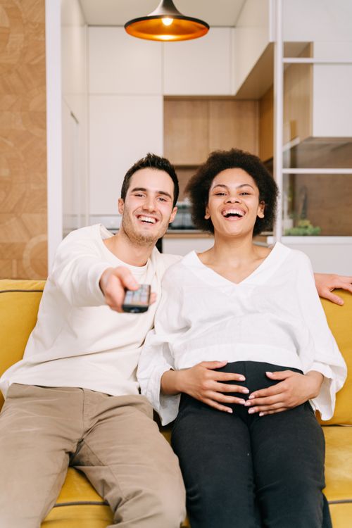 Free Smiling couple sitting on sofa enjoying time together, with remote control, expecting mother. Stock Photo