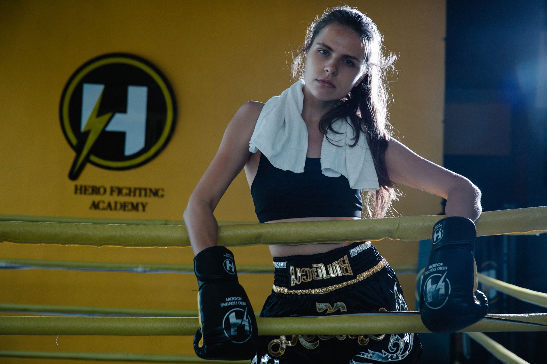 Young woman boxer at Hero Fighting Academy leaning on ring ropes with boxing gloves and towel.