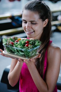 Smiling woman in a pink top holding a bowl of fresh salad with cherry tomatoes and greens indoors.