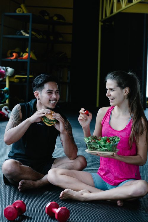 Free A couple laughs while sharing a salad and burger sitting on a gym floor. Stock Photo