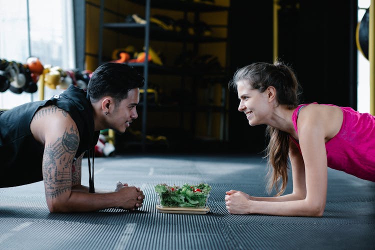 Man And Woman At The Gym Doing Planks While Looking At Each Other And Smiling 