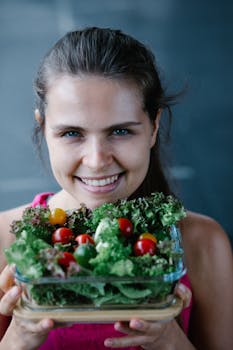 A happy woman in a pink top holds a healthy vegetable salad, showcasing fresh greens and cherry tomatoes.