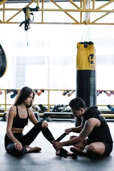 Trainer helps woman with wraps in a gym setting, surrounded by punching bags and sports equipment.