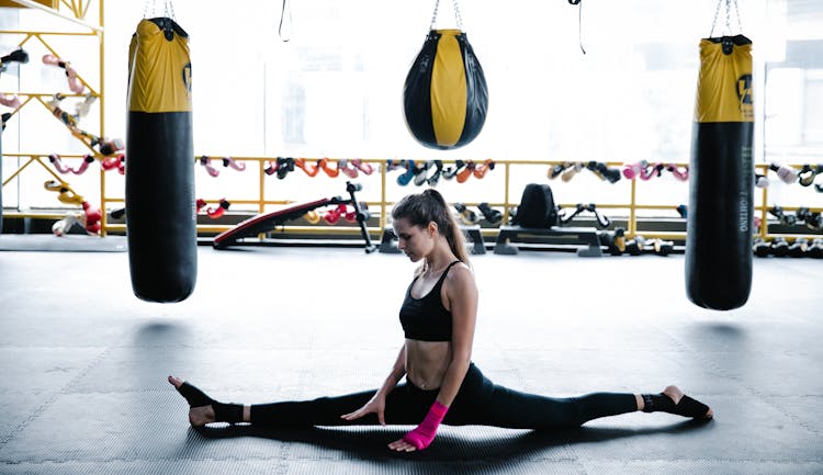 Woman In Black Tank Top And Leggings Doing Split Beside Black And Yellow Punching Bag