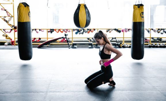 Woman kneeling on gym mat prepping with boxing equipment for workout.