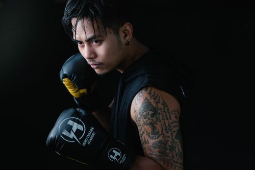 Free Focused boxer with tattoos and gloves poised for action in a dimly lit studio setting. Stock Photo