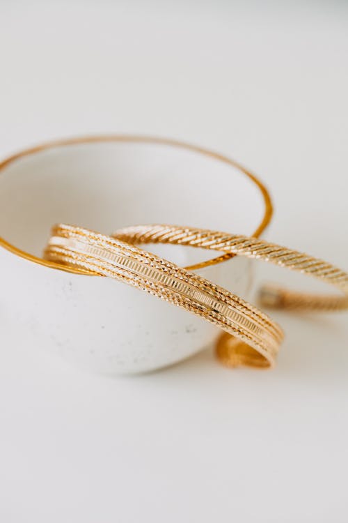 From above of shiny golden bracelets placed in porcelain cup on white table in daylight