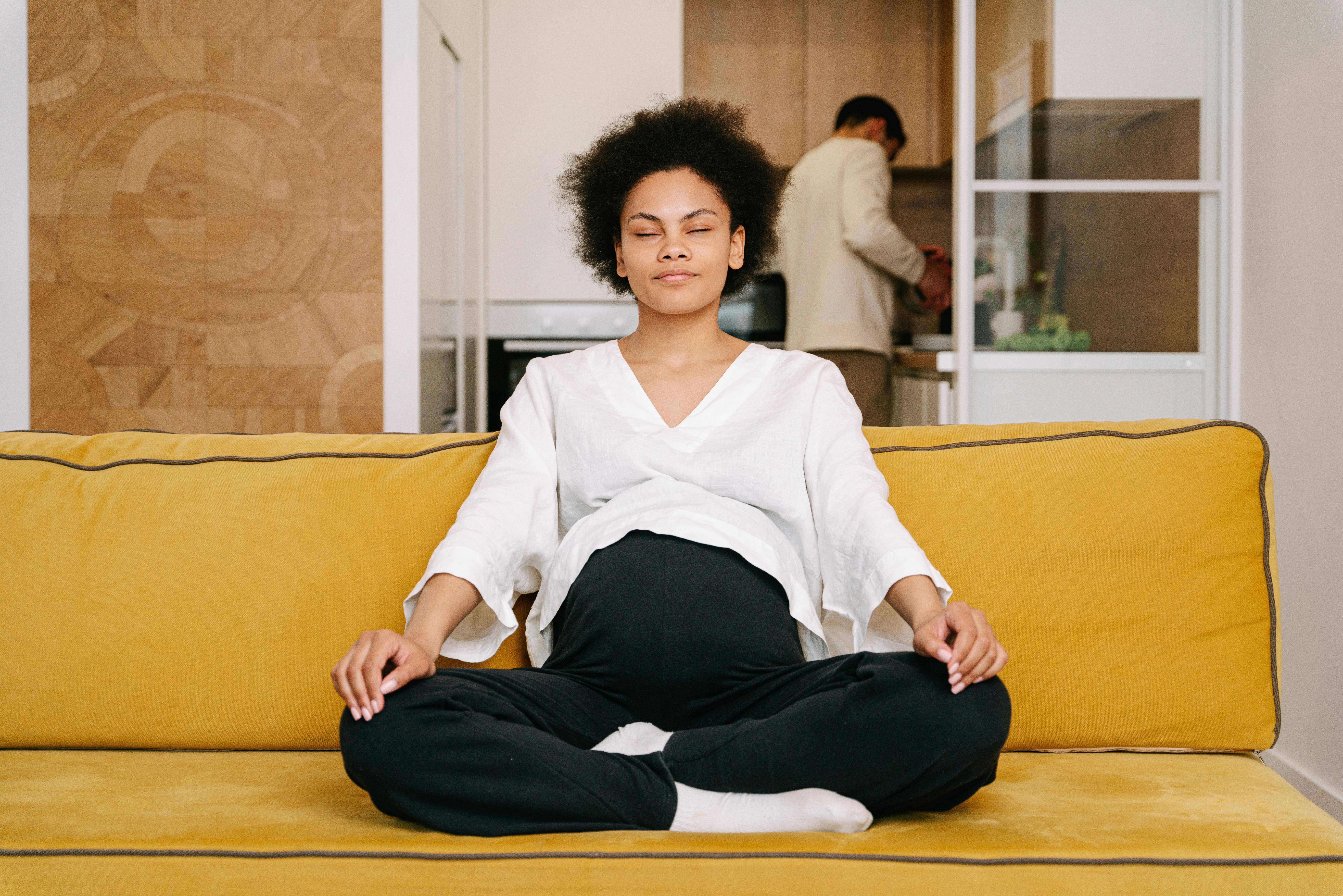 Expectant woman meditating on a yellow couch in a cozy home setting, emphasizing relaxation and self-care.
