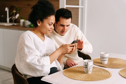 Free Happy couple sitting in kitchen browsing online shopping on smartphone, sharing moments together. Stock Photo