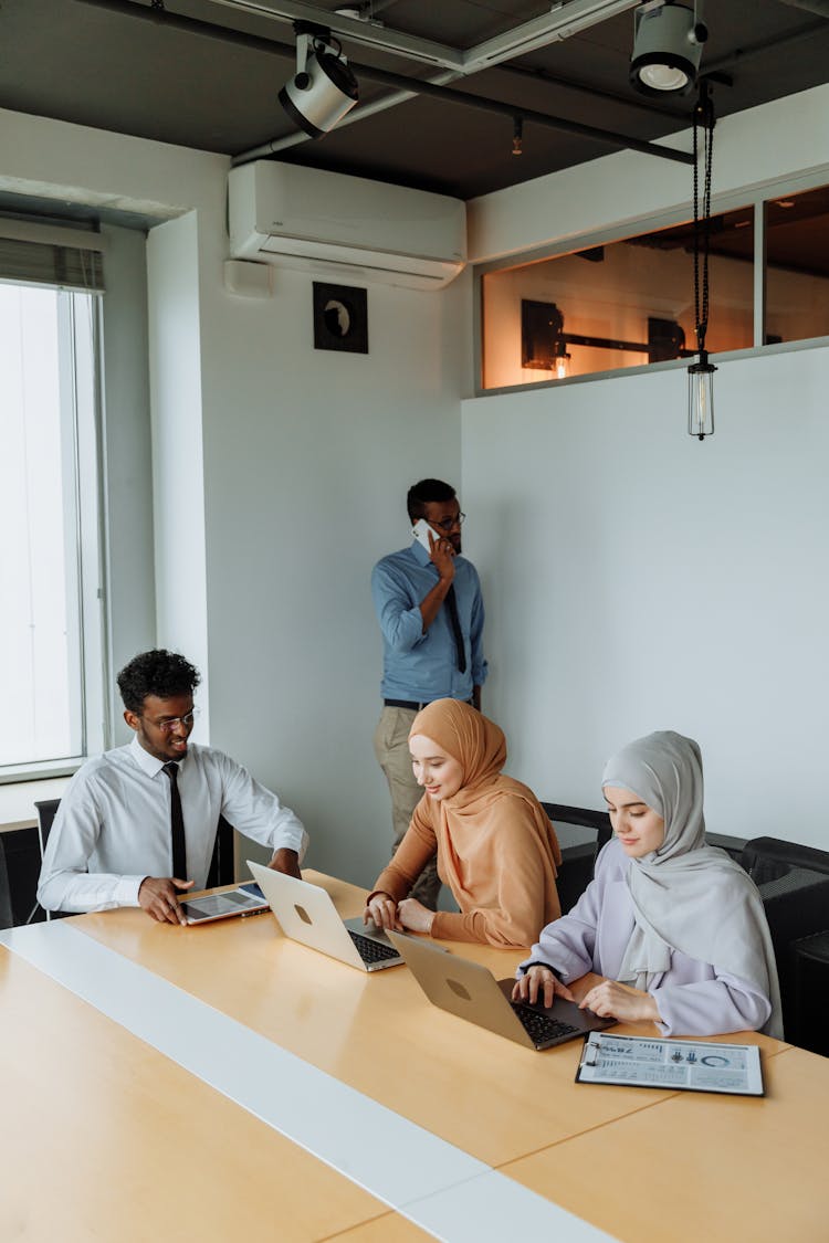 A Man In Blue Long Sleeves Talking On The Phone Near His Colleagues Working On The Table Using Laptop