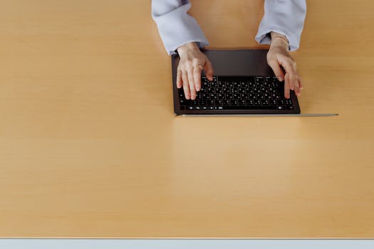 Top view of hands typing on a laptop placed on a wooden desk, emphasizing technology and workspace.