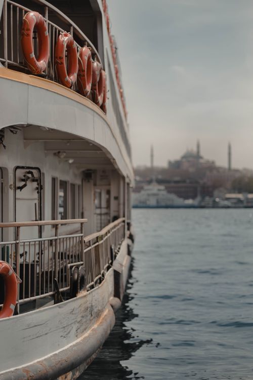 Free Side of modern white ship with lifebuoys floating on rippling water of sea near aged city on cloudy weather Stock Photo