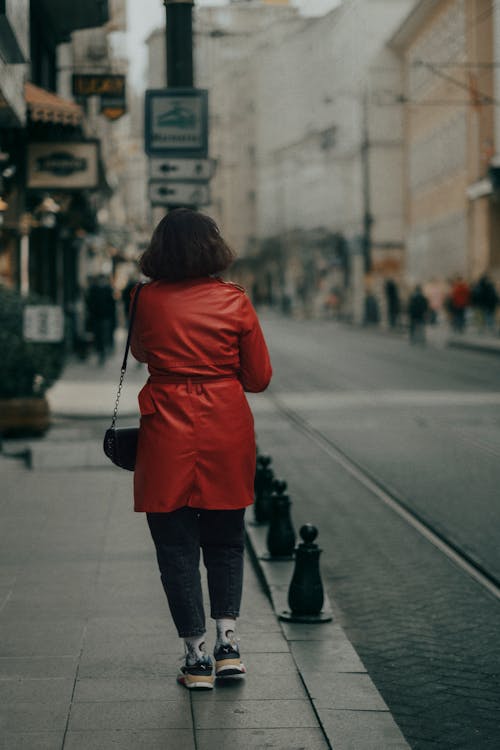 Back view of anonymous female with short dark hair and handbag wearing red coat strolling on pavement near road in city street in daylight