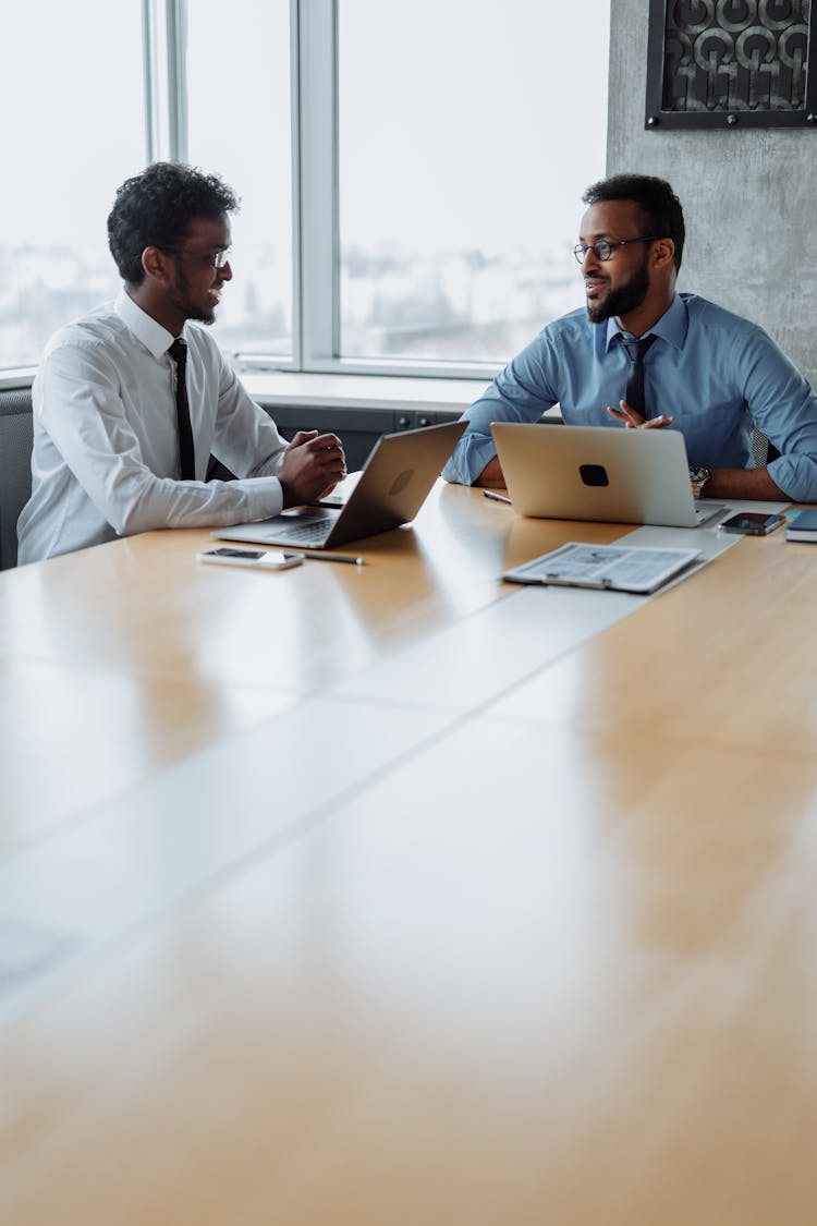 Two Men In Shirts Sitting At Table With Laptops And Talking