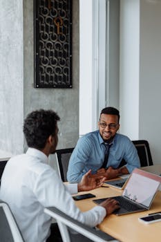 Two men in business attire discussing work at an office table with laptops.