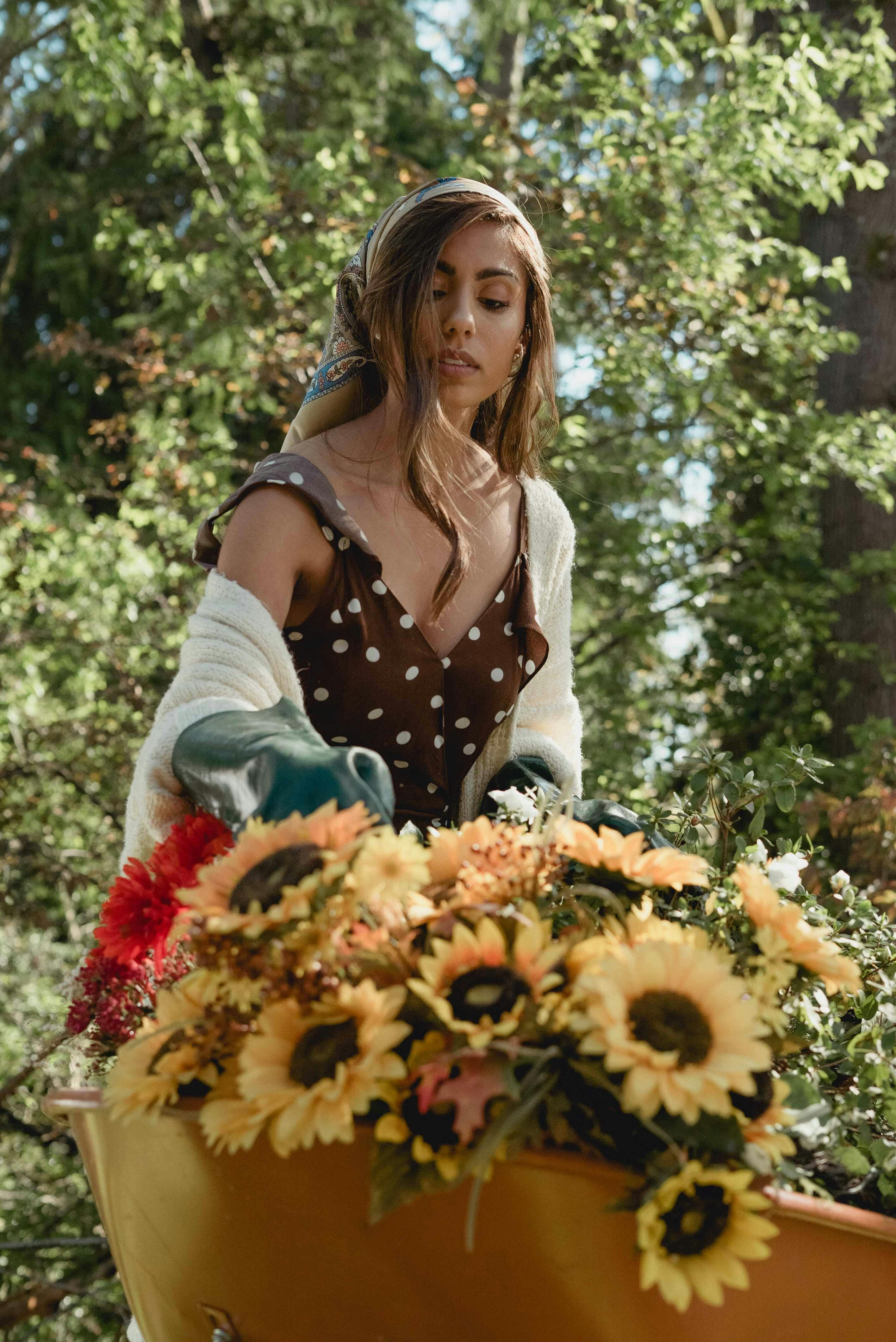 A woman gathers sunflowers in a garden, wearing a polka dot dress and sweater.