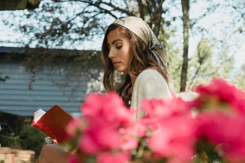 Free Young woman reading a book outdoors, surrounded by vibrant flowers, wearing a colorful scarf. Stock Photo