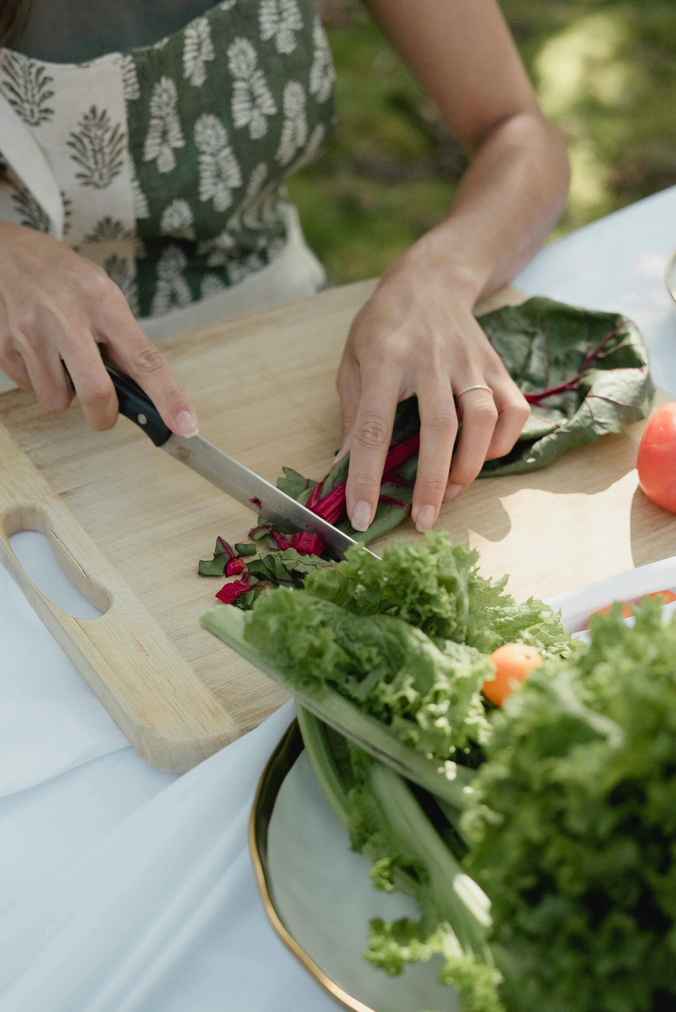 A person slicing swiss chard on a chopping board outdoors, surrounded by fresh vegetables.
