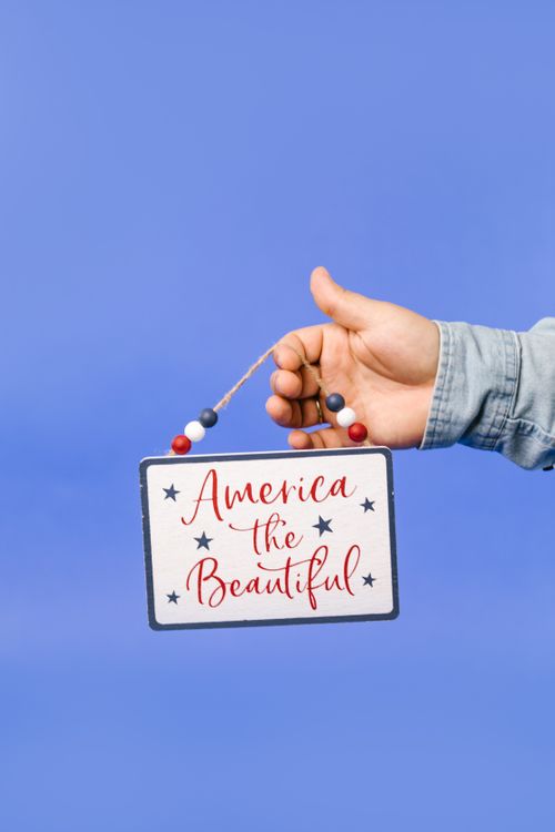 Free A hand holding an 'America the Beautiful' sign against a blue background, symbolizing patriotism. Stock Photo