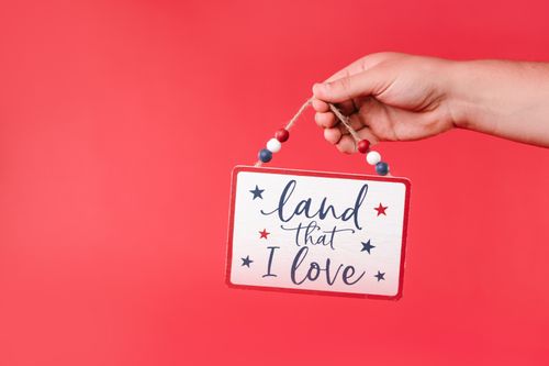 Free Hand holding a patriotic sign with stars and text 'land that I love' on a red background. Stock Photo