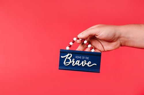 Free Hand holding a patriotic sign with 'Home of the Brave' against a red backdrop. Stock Photo