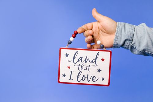 Free A hand holding a patriotic sign reading 'Land that I love' on a blue background. Stock Photo