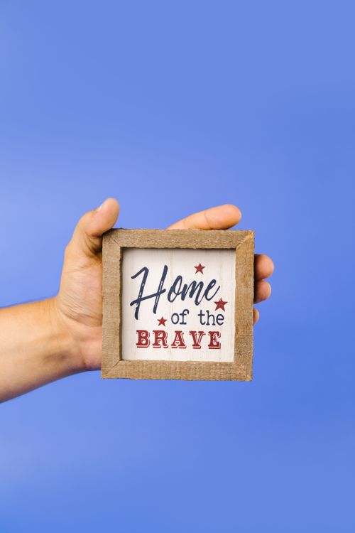Free A hand holds a wooden frame with 'Home of the Brave' text on a blue background. Stock Photo