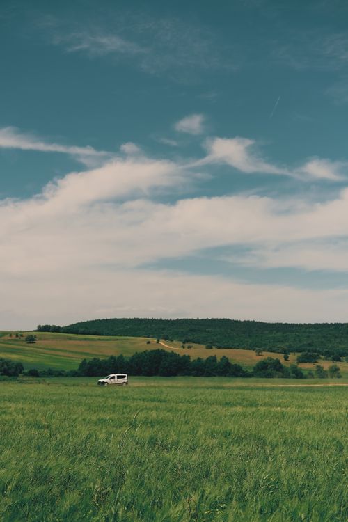 Free Green grassy field near vehicle and hills with trees under blue cloudy sky in daylight in nature Stock Photo
