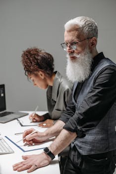 An African American woman and Caucasian man collaborate at a desk, working on computers and writing notes.
