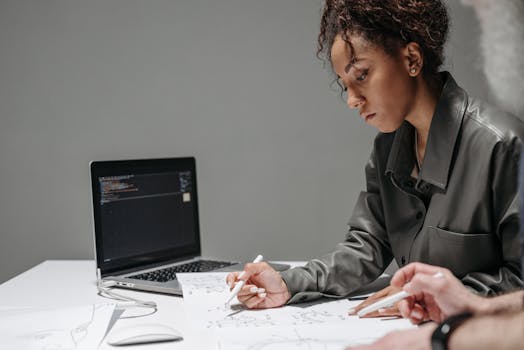 Young designer working on sketches with a laptop beside her, embodying creativity and focus.