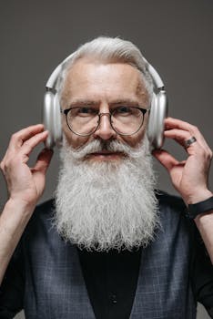 Senior man with white beard wearing wireless headphones and glasses, enjoying music indoors.