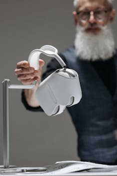 Senior adult man with a beard demonstrates a VR headset on a sleek stand indoors.