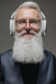 Close-up portrait of a senior man with a beard, wearing headphones and glasses, smiling.