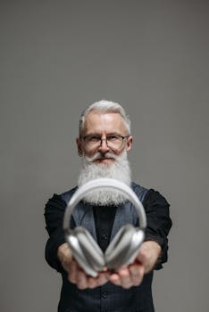 Senior man with beard presenting headphones in a studio setting with grey background.