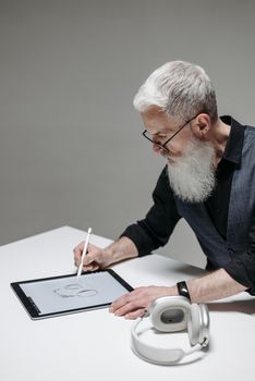 Elderly man sketching with digital pen on a tablet at a desk, showcasing modern technology use.