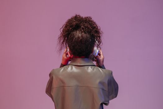 A woman with curly hair wearing a leather jacket and headphones, against a purple background.