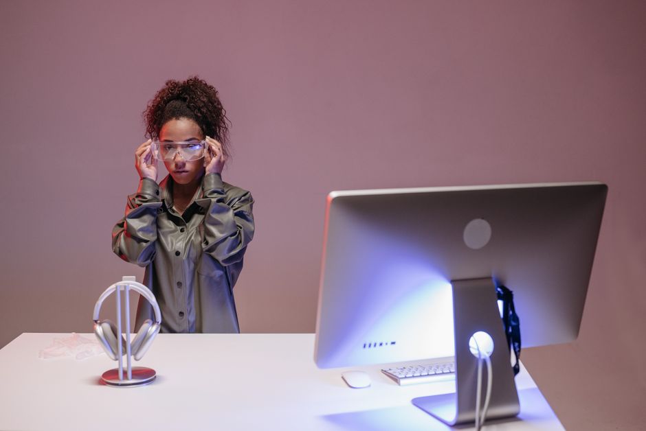 Black woman adjusting VR goggles at desk, modern workspace with tech equipment.