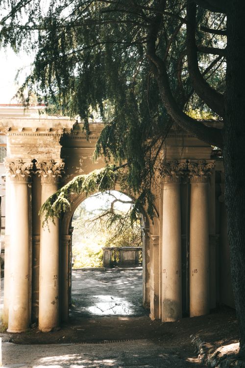 Free A sunlit classical archway surrounded by trees in a serene park environment. Stock Photo