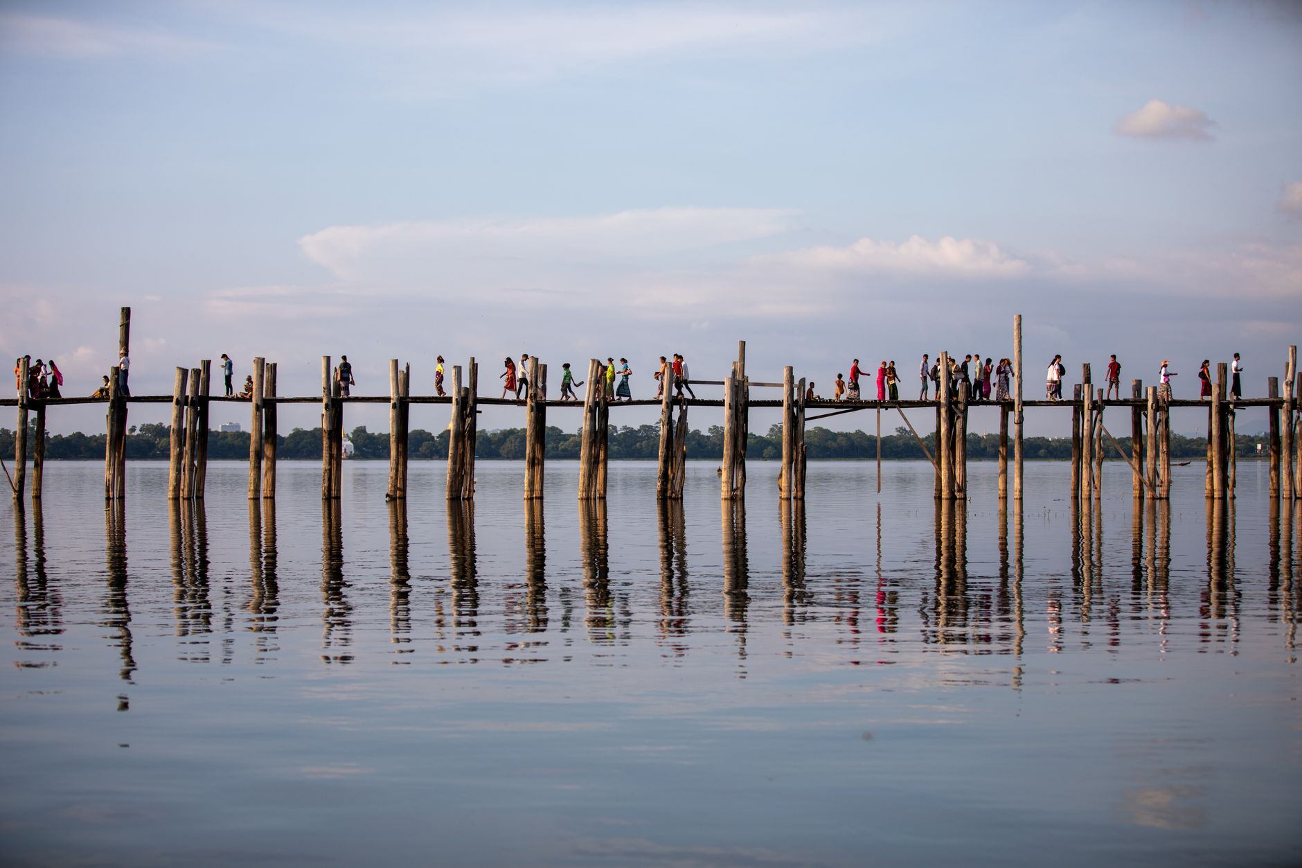 People walking on the iconic U Bein Bridge, with a serene water reflection in Amarapura, Myanmar.