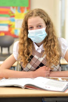 Young girl with mask sitting at desk with an open book in a classroom.