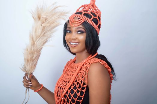 Portrait of a smiling woman in traditional African attire with a beaded crown and holding a pampas grass.