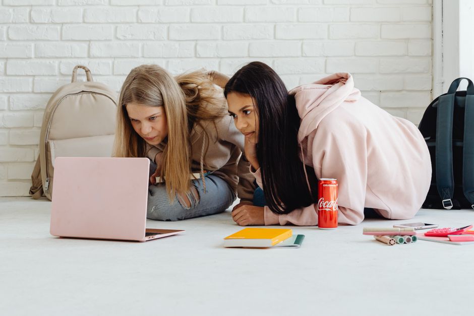 Two teenage girls studying indoors using a laptop, surrounded by backpacks and stationery.