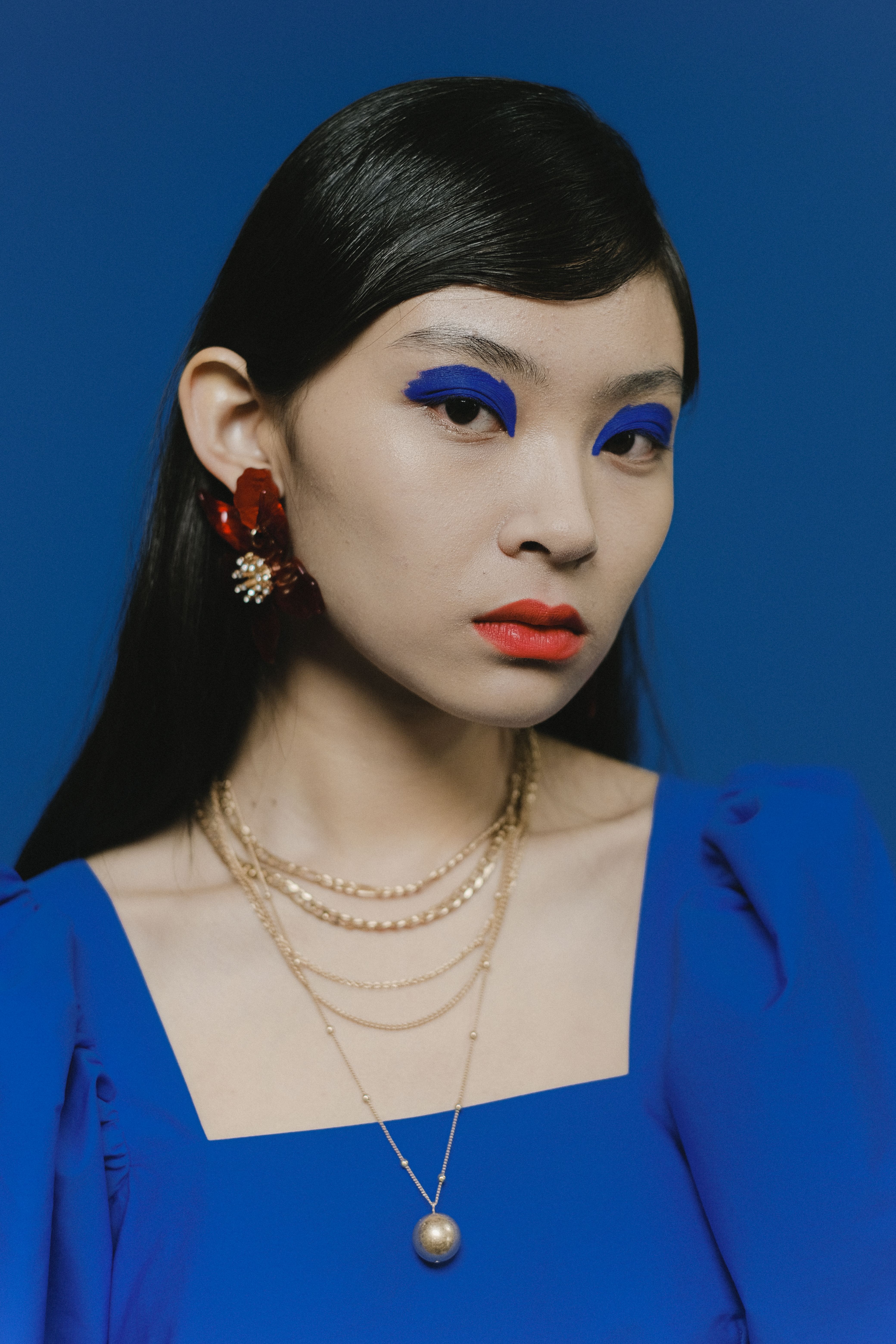 Portrait of a fashionable Asian woman with vibrant blue makeup and jewelry against a blue backdrop.